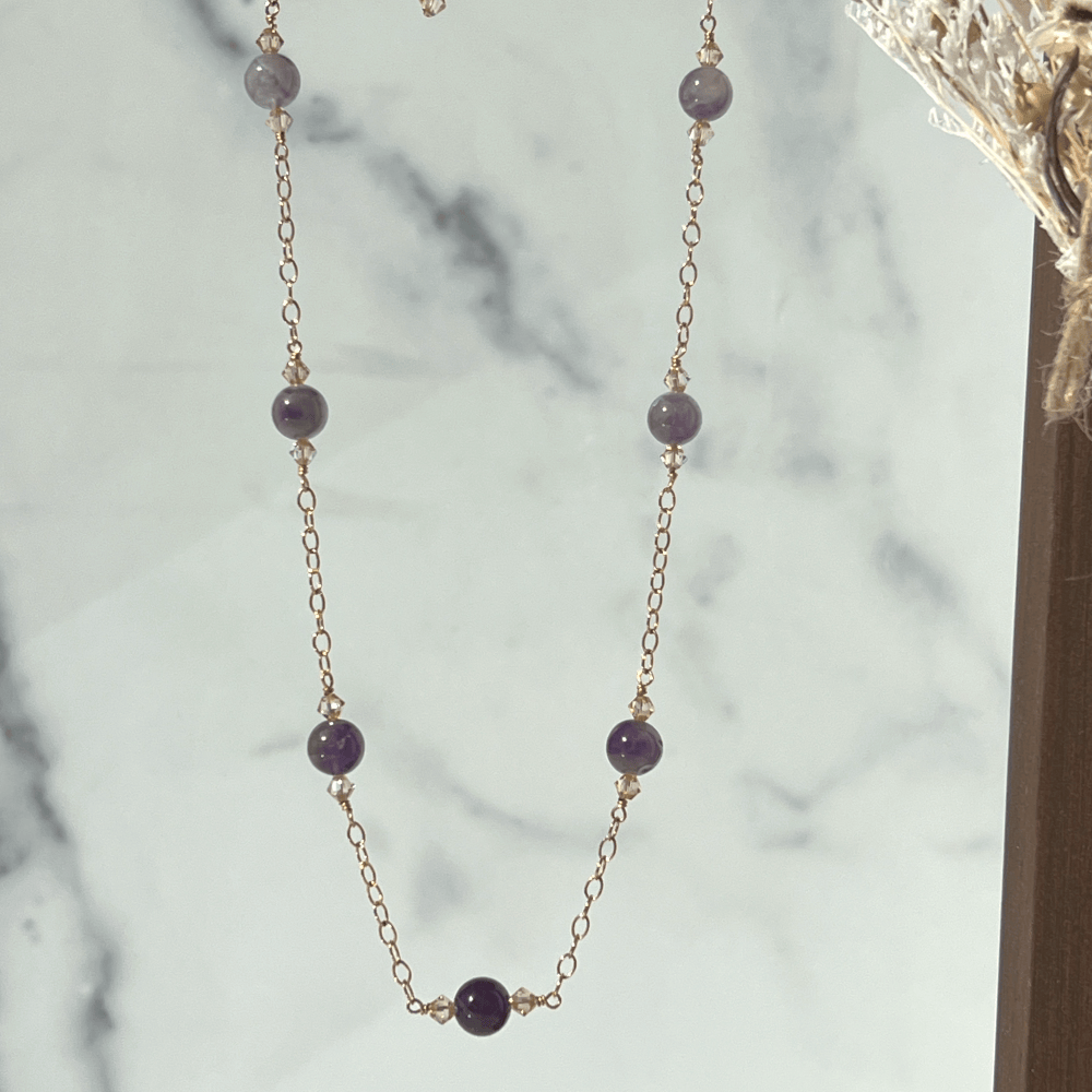 Make a glamorous statement with this stunning ombré-style necklace featuring natural Amethyst gems, Swarovski crystal golden shadow beads, and a 14Kt rose-gold filled chain. The unique design adds a flowing ombré pattern that captivates the eye. Perfect to add a touch of elegance to your everyday wardrobe. Modeled with matching earrings and a single pearl on a rose gold chain choker.