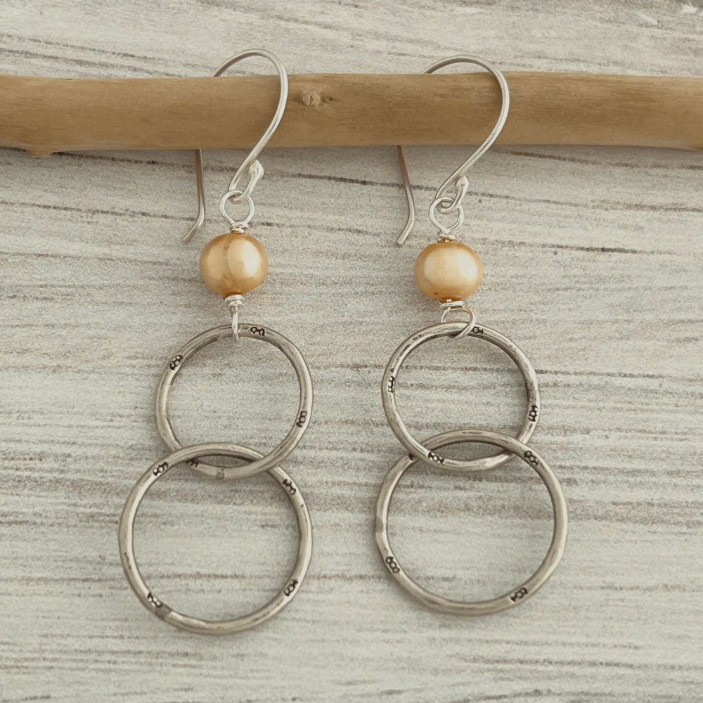 These artisan earrings bring together the richness of heritage silverwork with the soft radiance of apricot pearls. Two interlocking hoops in high-purity Hilltribes silver are paired with luminous apricot freshwater pearls, creating a design that feels both rooted and refined.