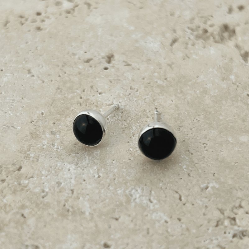 Close-up of handcrafted black onyx stud earrings set in polished sterling silver bezels on neutral stone background.