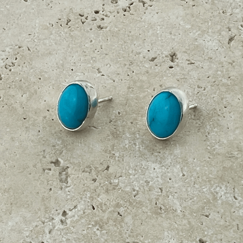Close-up of oval Kingman Turquoise stud earrings set in polished sterling silver bezels on a neutral stone background.