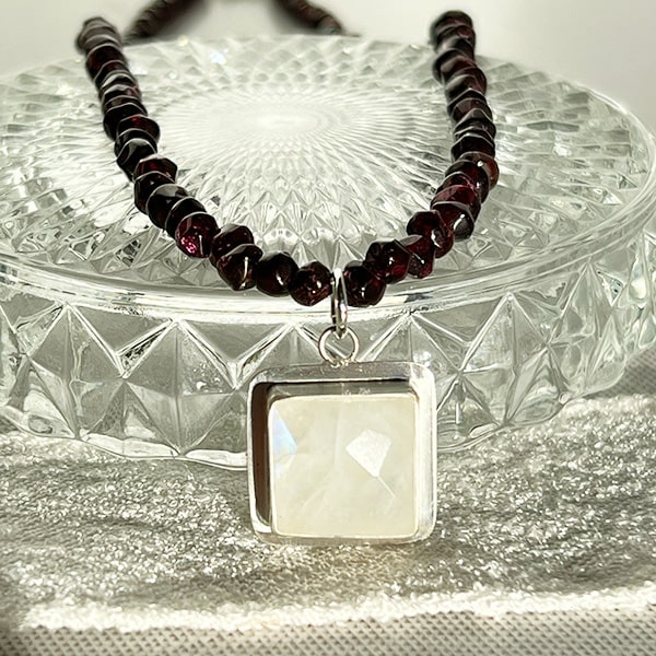 Discover the ethereal allure of this Rainbow Moonstone, a gem with a mesmerizing play of colors that dance and shimmer as if capturing a slice of the northern lights. Complementing the moonstone's ethereal charm is a necklace of garnets, their deep crimson hue symbolizing passion and vitality. Handcrafted in CA.