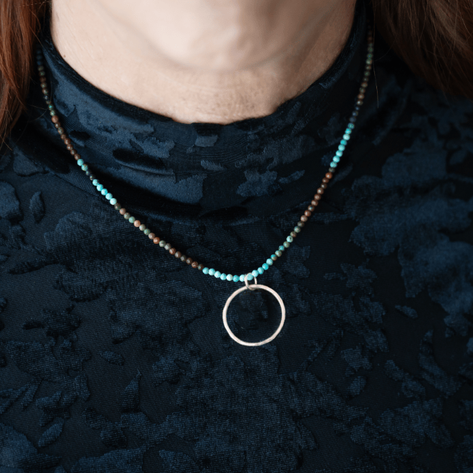 Turquoise beaded necklace with a open-circle sterling silver pendant worn by a person with a blace blouse. 