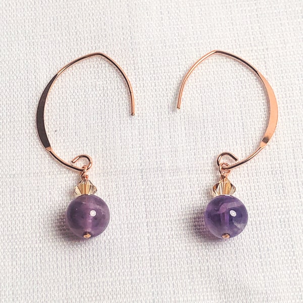 These elegant Amethyst earrings from our collection promise to make a chic statement wherever you go. The flat V-shaped half-hoop ear wire is crafted from 14Kt Rose Gold-filled metal, and each earring is adorned with a shimmery golden Swarovski crystal bead.